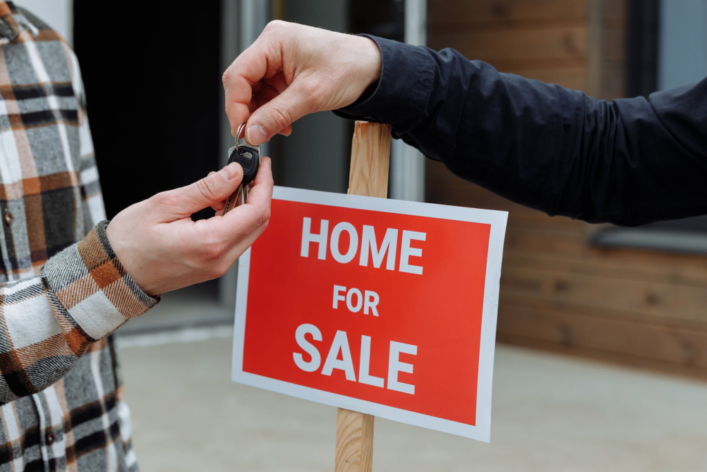 A person handing out the keys in front of a "Home for Sale" sign
