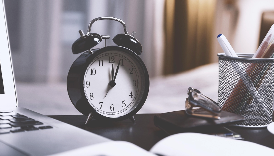 A table clock next to a computer and a pencil holder.