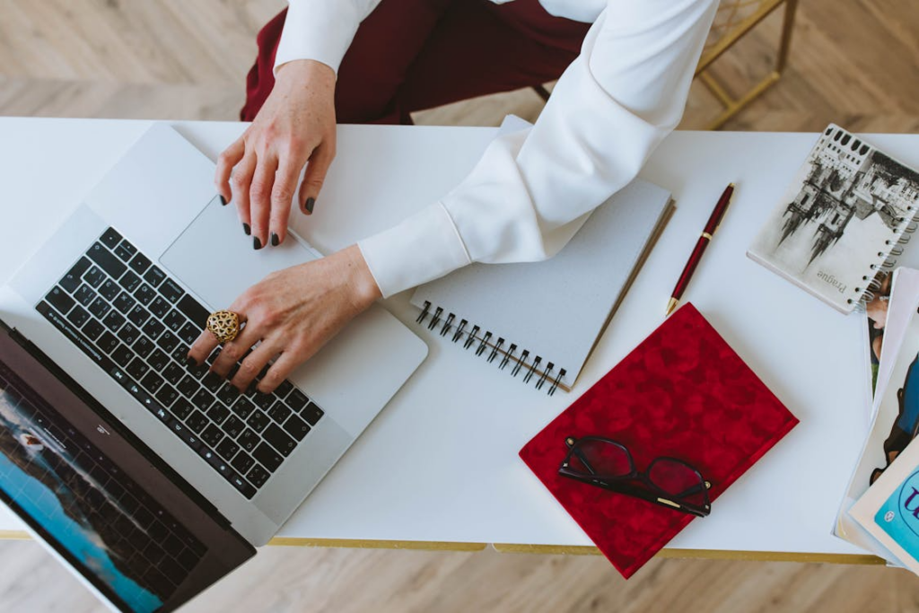 A person typing on a laptop at a desk with a notebook, pen, and glasses.