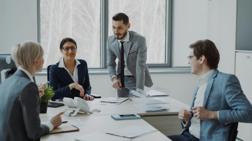 Four business professionals in a conference room discussion, with one man standing and presenting while three seated colleagues listen attentively.