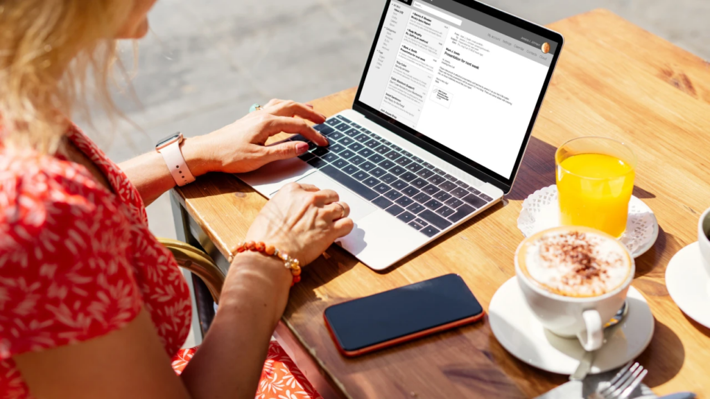 Woman checking email on a laptop at a café table with a cappuccino, orange juice, and smartphone.