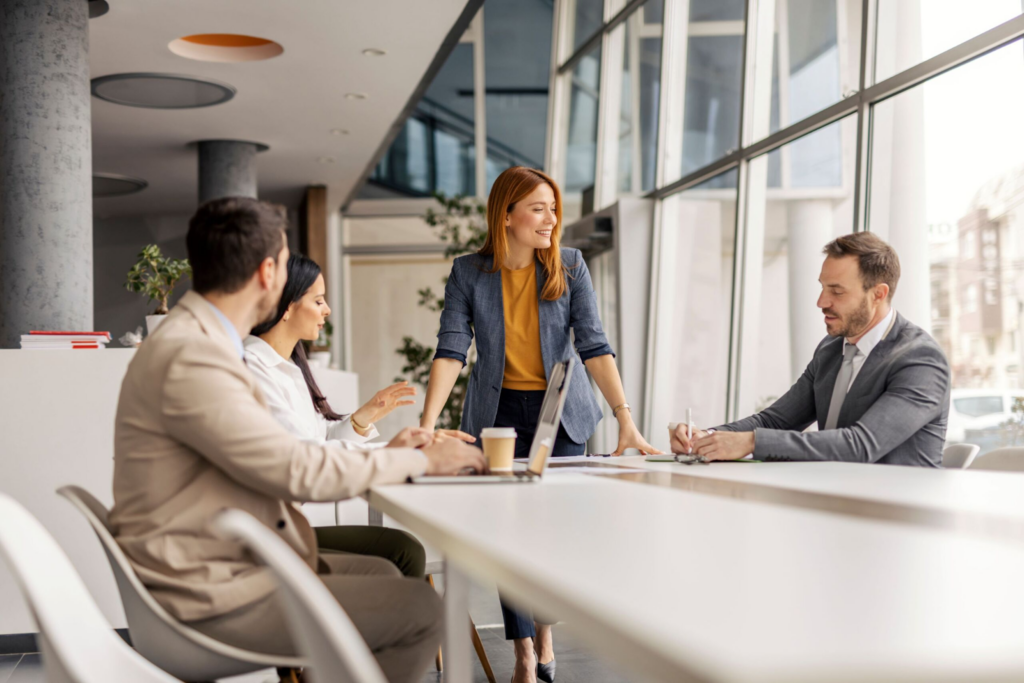 Team of four professionals in a business meeting at a bright, modern office with large windows.