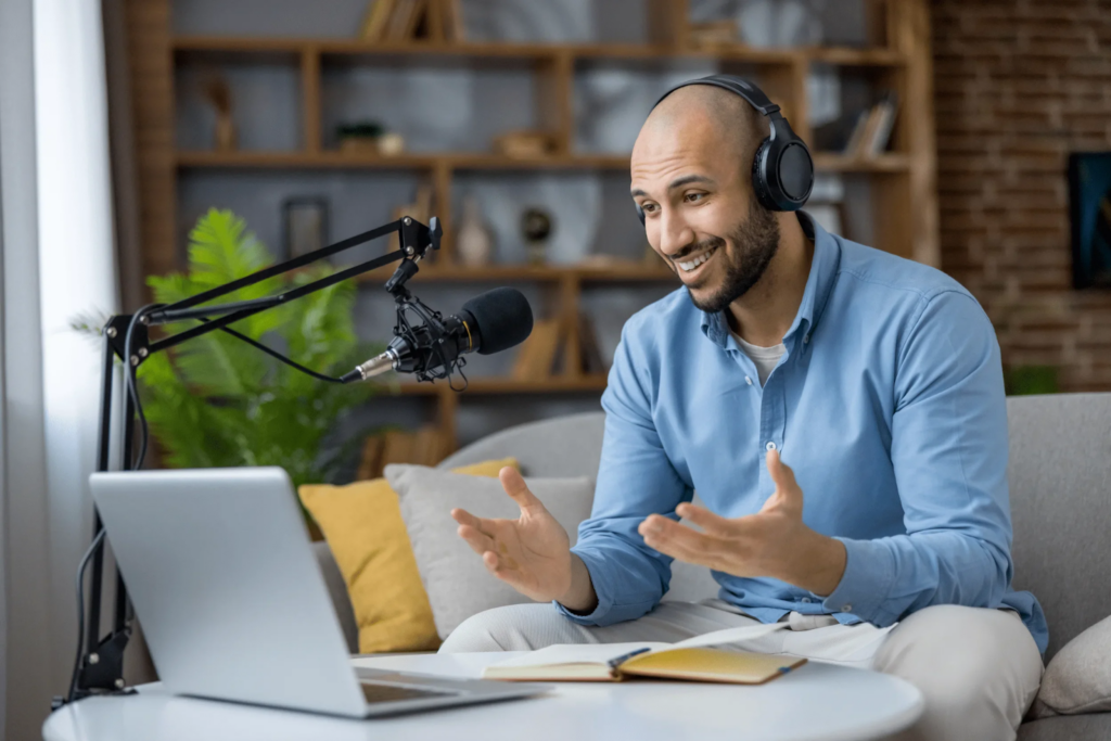 Man wearing headphones speaking into a boom microphone while recording a podcast from a cozy home studio setup with a laptop and notebook.