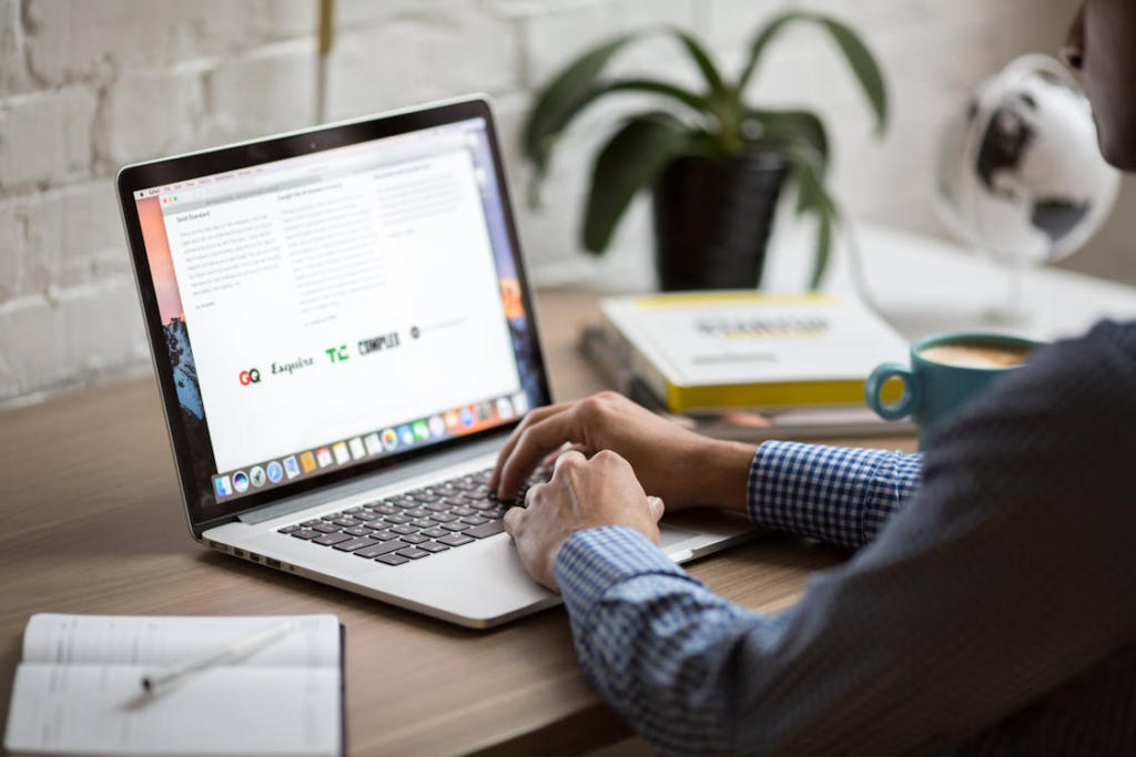 Person typing on a laptop displaying a structured document on a desk with a notebook nearby.