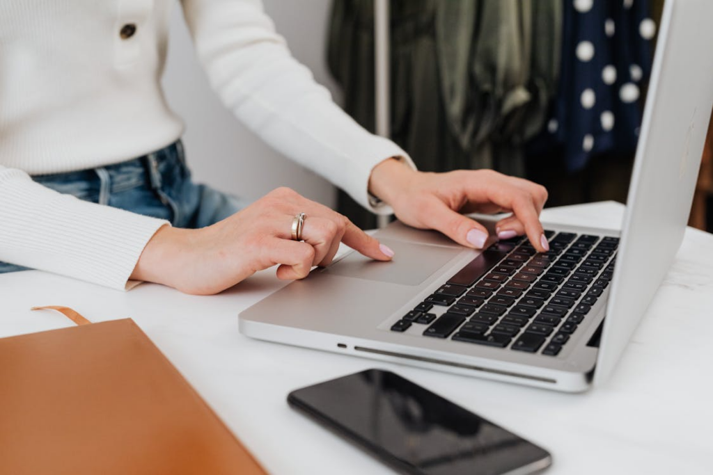 A person typing on a laptop at a white desk with a notebook and phone beside it.