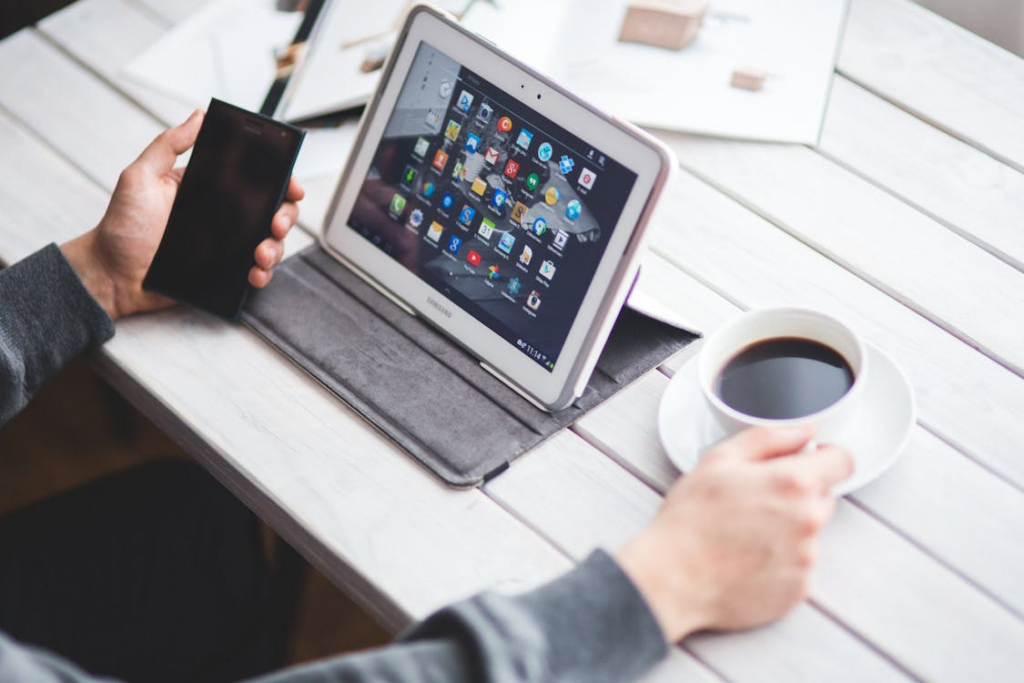 A person holding a phone and a cup of coffee with a tablet on the table showing various apps.