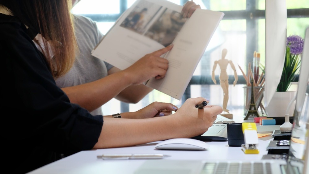 Two people reviewing printed materials at a creative workspace with a wooden mannequin figure, pens, and a computer monitor.