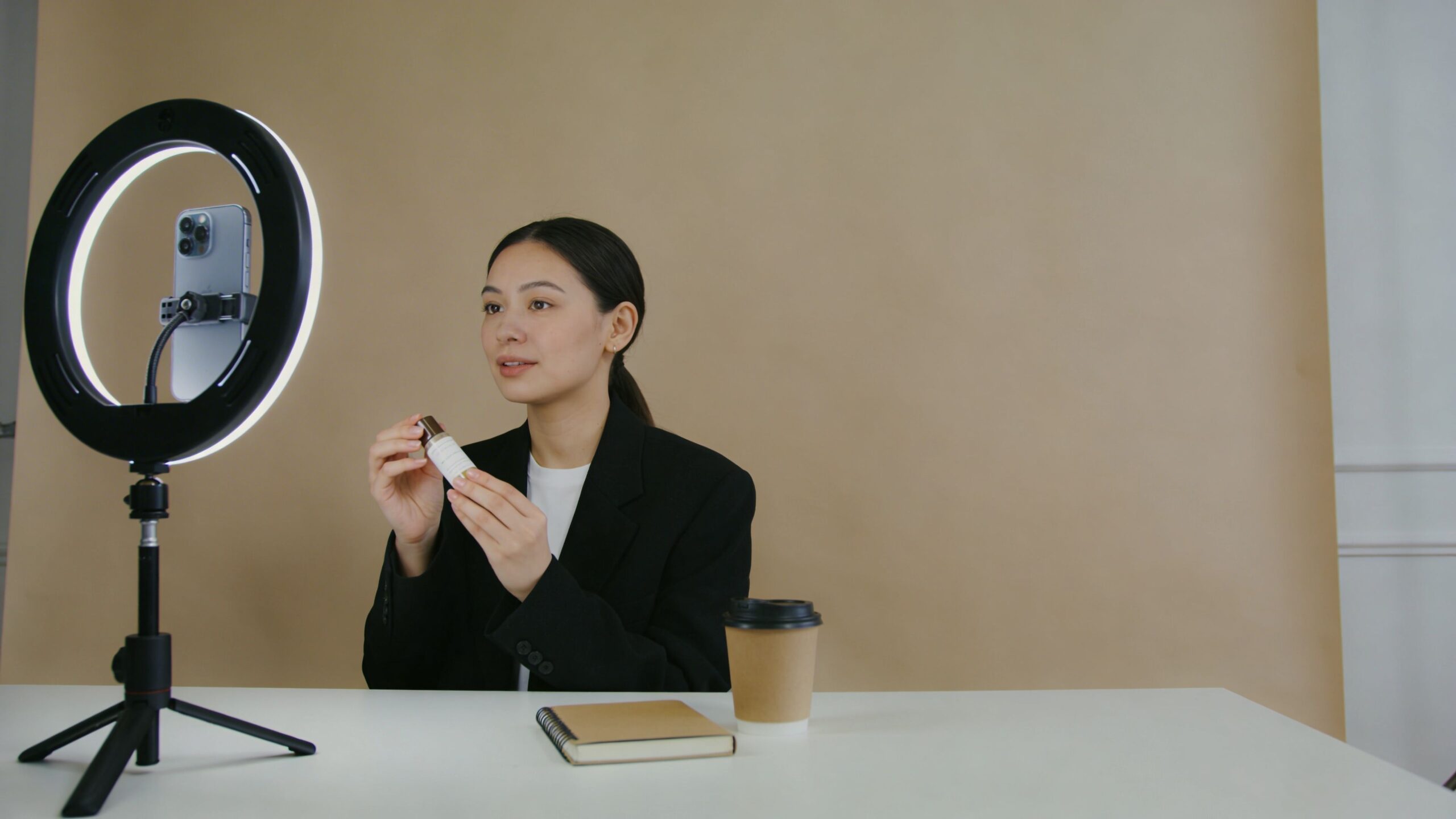 A young woman holding a skincare product while recording a video for her online audience using a ring light.