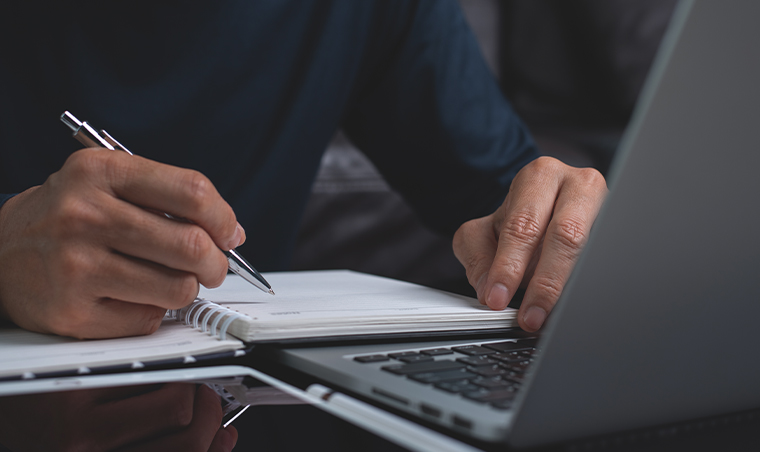 Person in a blue shirt handwriting notes beside a laptop while refining an AI-generated press release draft